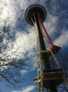 View of the Space Needle against a cloudy sky, with a large red object attached to its side, resembling an out-of-state investment in architectural marvels. Bare tree branches are in the foreground.