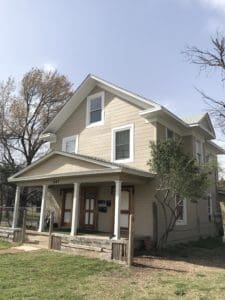 A charming two-story beige house with white trim and a porch, nestled amid lush trees, exemplifies the beauty of Oklahoma City real estate.