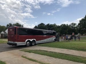 A red and white tour bus parked on a suburban street in Oklahoma City, with a group of people gathered nearby and a cloudy sky overhead, adds charm to the local real estate scene.