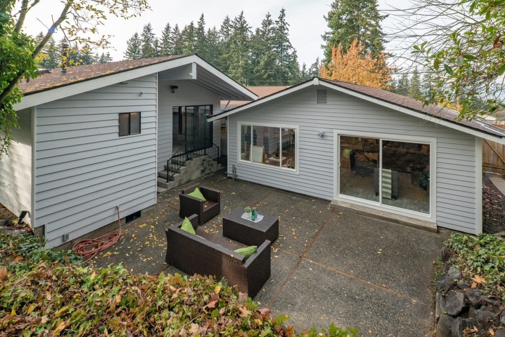 Aerial view of a backyard with a concrete patio, outdoor seating, and two white houses surrounded by trees—a perfect setting for flipping houses with its spacious layout and serene environment.