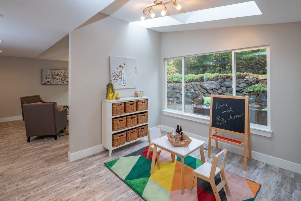 A playroom designed with a house-flipping flair, featuring a colorful rug, small table and chairs, chalkboard, storage cabinet with baskets, and a large window overlooking the garden.