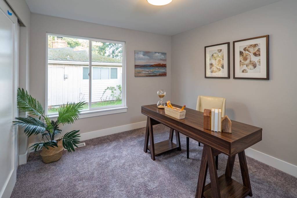 A home office with a wooden desk, a beige chair, books, and a decorative hourglass evokes the perfect workspace for flipping houses. Two framed artworks add charm to the walls, while a potted plant sits near a window overlooking a yard.
