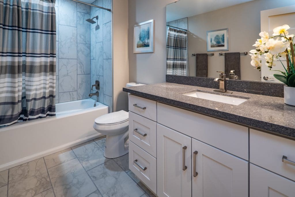 A modern bathroom perfect for flipping houses, featuring a white bathtub, striped shower curtain, large mirror, granite countertop, and sleek white cabinets. Decorative touches include a vase of flowers and elegant wall art.