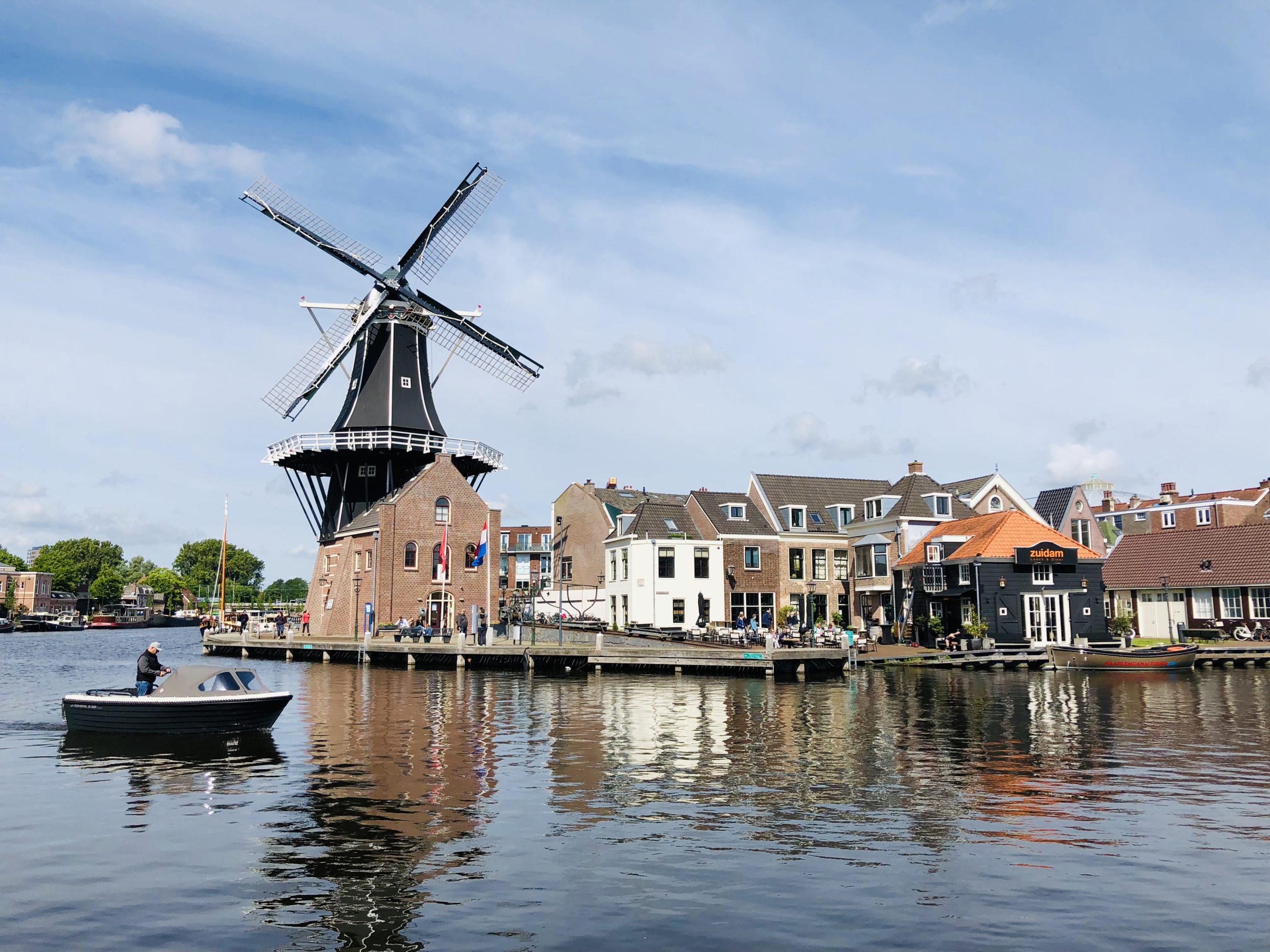 A windmill by a canal with boats and traditional buildings under a cloudy blue sky sets the scene, reminiscent of finding the right time to start investing in real estate—timeless and picturesque.