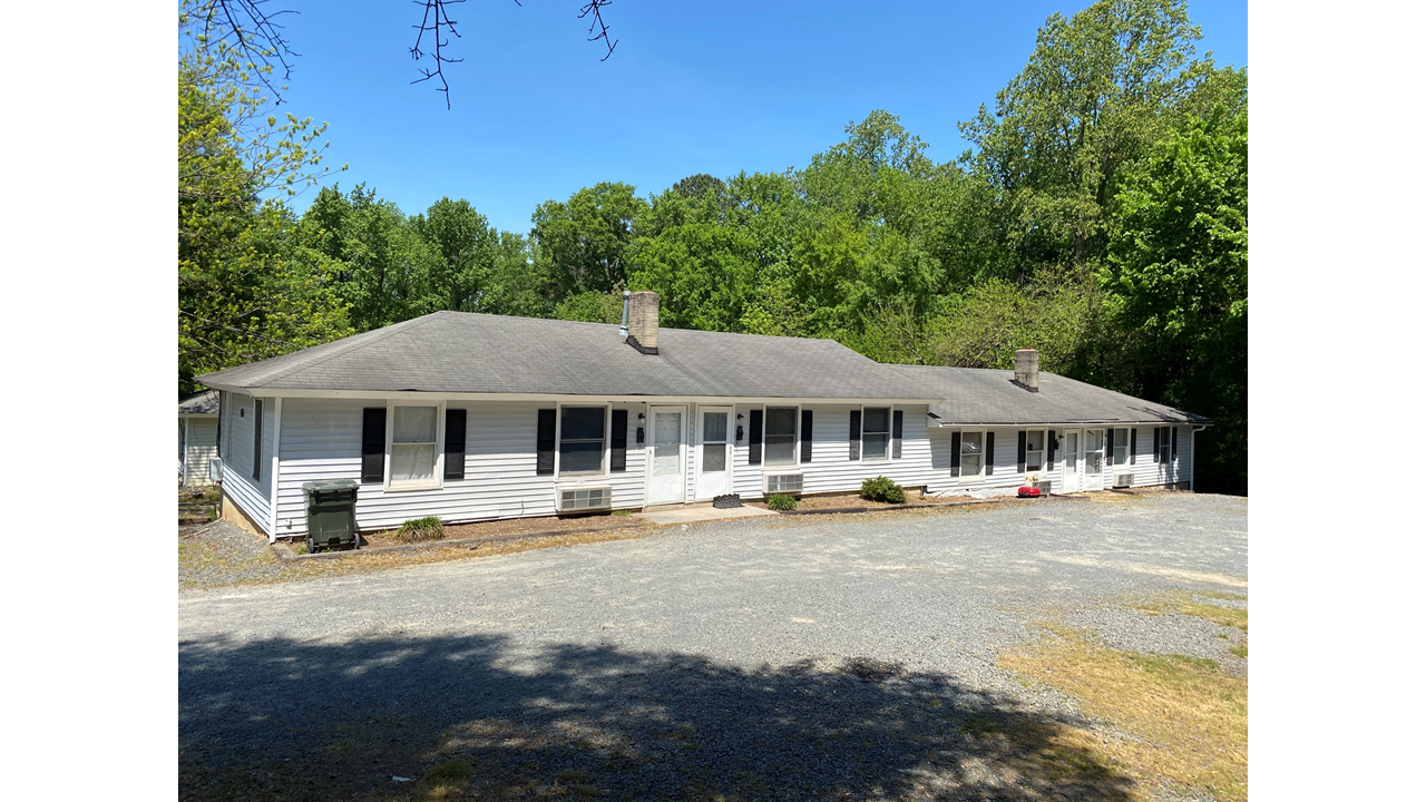A long, single-story white house with a gray roof embodies financial freedom through real estate investing. Its large windows and two chimneys complement the backdrop of green trees and a clear blue sky, while the gravel driveway invites you in.