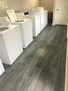 A bright laundry room features four white washing machines and dryers perfectly lined up along a wall on a gray tiled floor, creating an efficient space for all your laundry needs. A closed door is visible at the end of the room, adding to its clean and organized appeal.