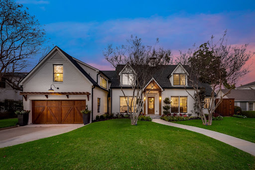 A two-story house with a brick exterior, wooden garage doors, and a well-kept lawn stands proudly against the vibrant hues of a sunset sky, tempting potential purchases with its charm.