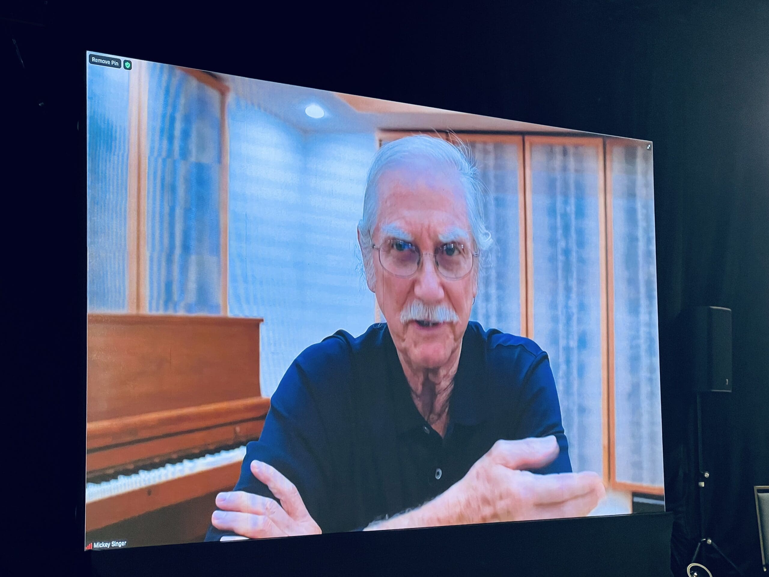 An older man with a mustache is speaking during a video call, with a piano in the background.