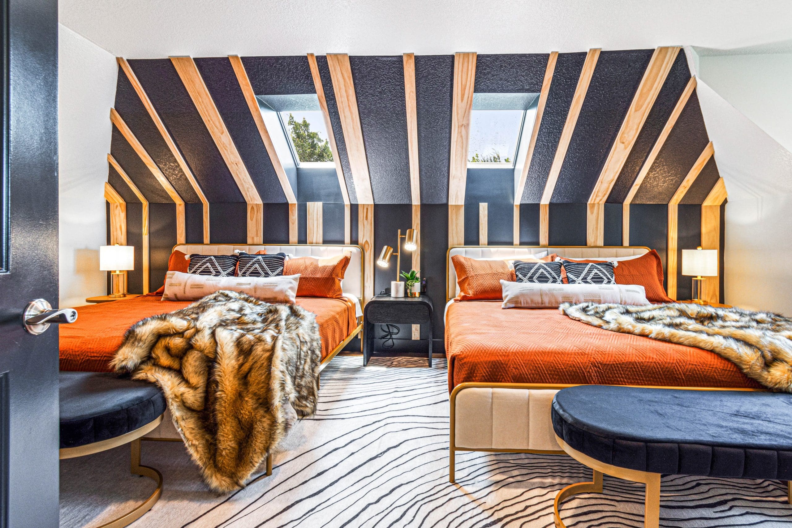 A bedroom designed to add value to a property features two beds with orange bedding and fur throws, complemented by striped walls. Skylights illuminate the space, while a zebra-patterned rug adds flair. Two lamps flank a central nightstand adorned with plants.