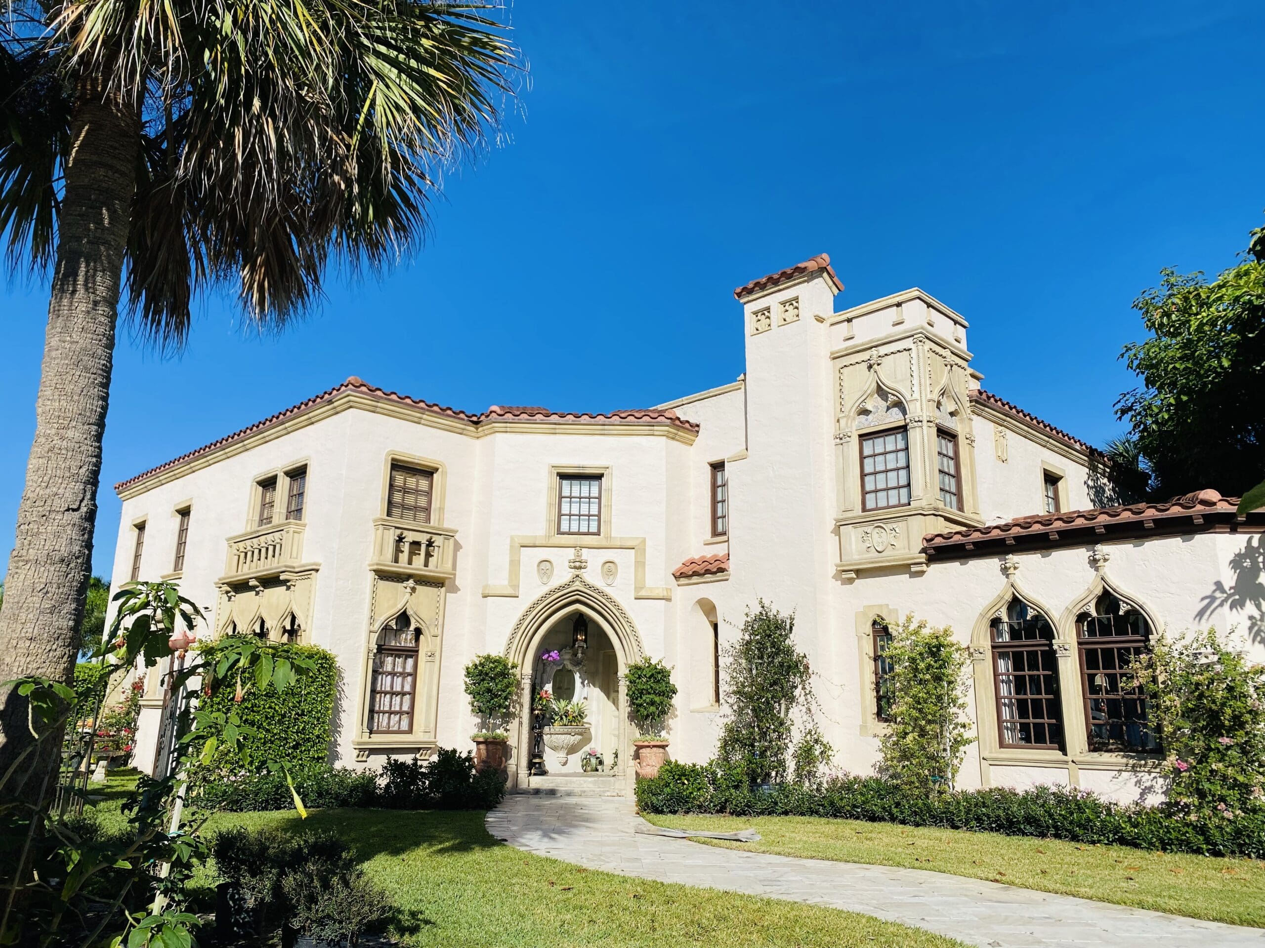A large, two-story house with beige stucco walls, red-tiled roof, arched windows, and a palm tree in the front under a clear blue sky.
