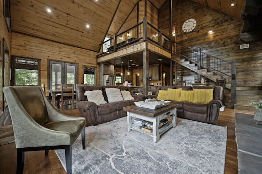 Best short-term rental markets Broken Bow OK Spacious rustic living room with wooden walls and ceiling, featuring leather sofas, a large rug, and a loft area with a clock. Ideal for short-term rental markets, the space includes a staircase and dining area visible in the background.