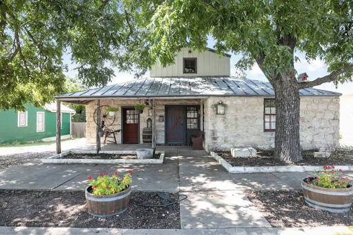Best short-term rental market Fredericksburg, TX A rustic stone house with a metal roof, surrounded by large trees, offers charm in the short-term rental market. Two planters with flowers grace the entrance, inviting guests to enjoy a serene escape.