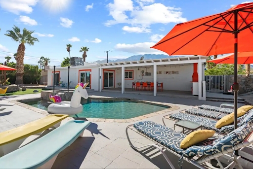 Best short-term rental market Greater Palm Springs, CA A backyard oasis ideal for short-term rental markets features lounge chairs, a unicorn float, and vibrant orange umbrellas. A patio with a long dining table rests under a white pergola, offering guests views of palm trees and mountains.