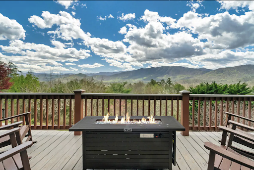 Best short-term rental markets Smoky Mountains, TN A wooden deck with chairs and a rectangular fire pit table overlooks a scenic mountain view under a partly cloudy sky, offering the perfect escape in the thriving short-term rental markets.