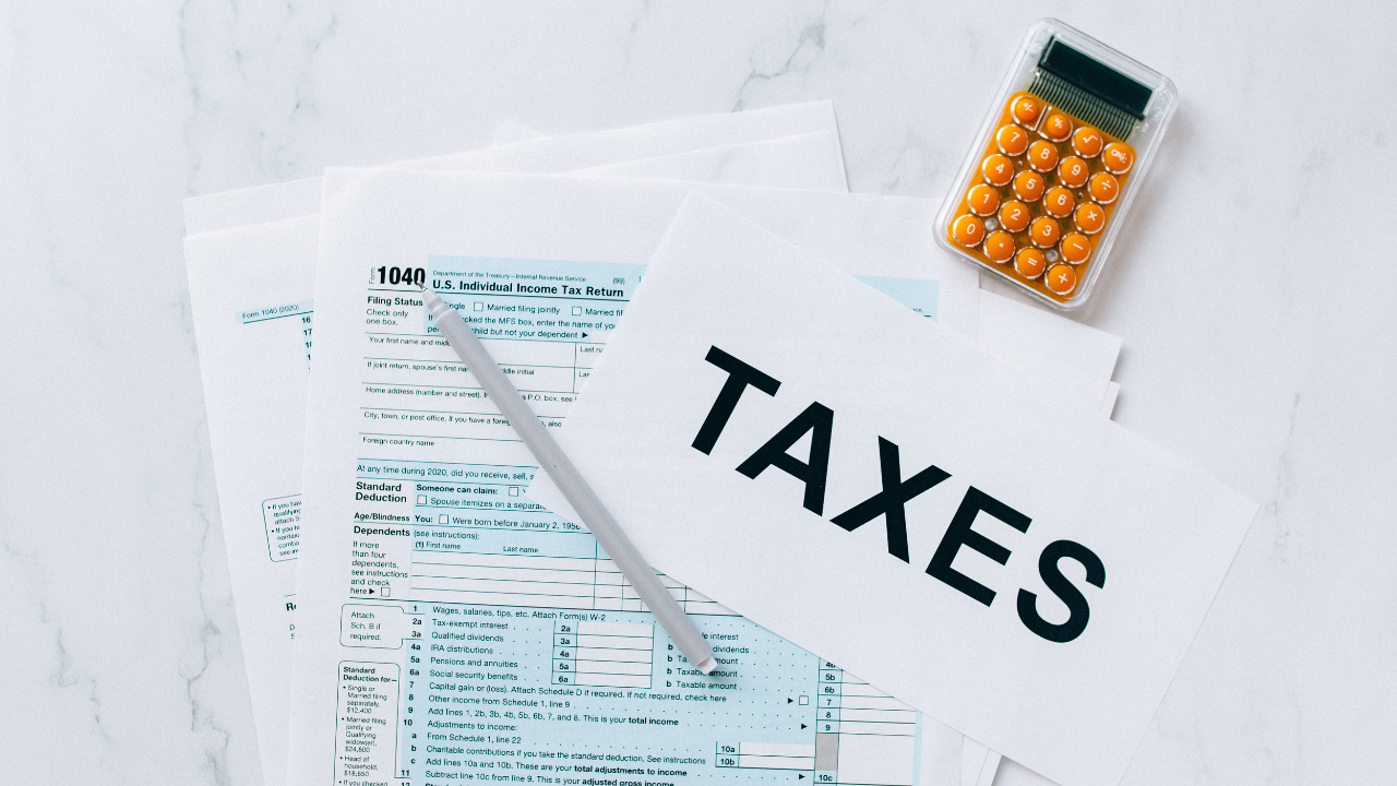 Tax forms, a pen, and a calculator with orange buttons are on a marble surface. One paper prominently displays the word "TAXES.