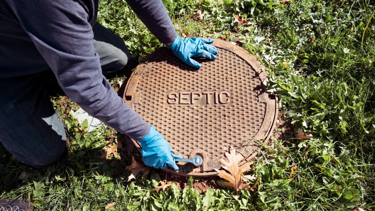 A person wearing blue gloves is kneeling on grass, using a tool to open a round septic tank cover with the word "SEPTIC" on it.