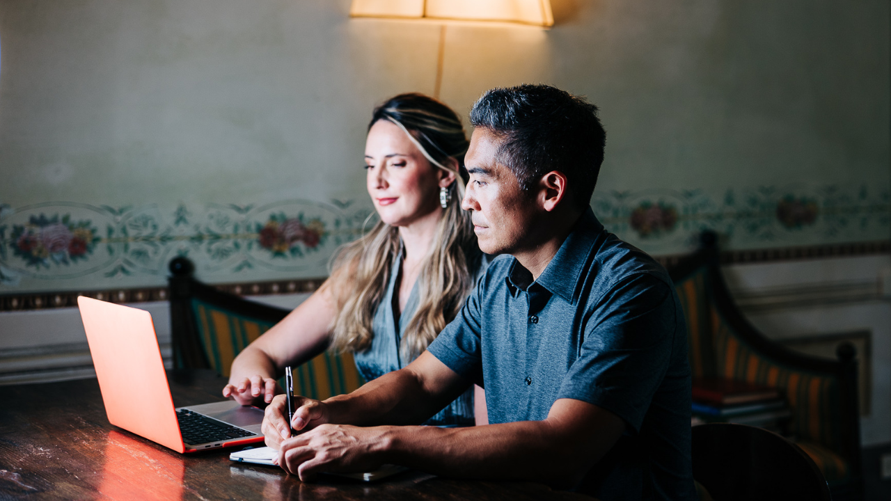Two people sitting at a wooden table, using a laptop. The room has vintage wallpaper and a soft light from a wall lamp.