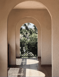 An arched hallway leads to a balcony with a tiled floor and a metal railing. Palm trees are visible in the background.