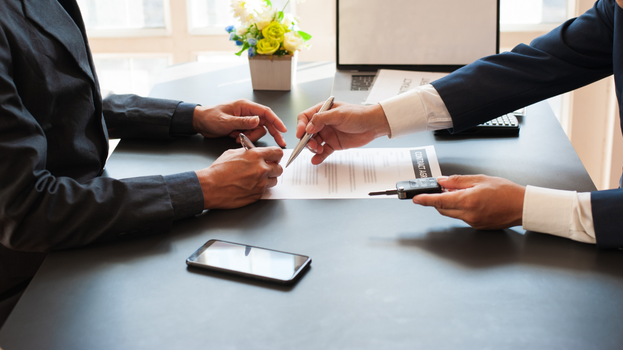 Two people in suits signing a document at a desk, with a smartphone and a small potted plant nearby.