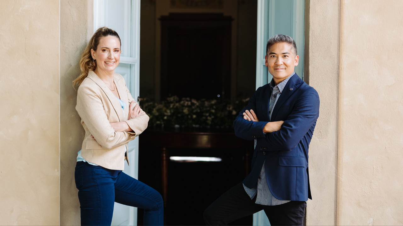 Two people stand with arms crossed, leaning against a doorway, discussing how to qualify for real estate professional status legally.