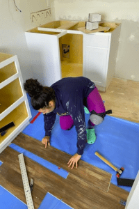 A person is installing wooden flooring in a room with cabinets. They are using tools and wearing knee pads and green Crocs. The floor is protected by a blue covering.