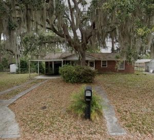 A brick house with a large tree draped in Spanish moss in the front yard. A concrete path leads to the front porch, and there's a black mailbox on the lawn.