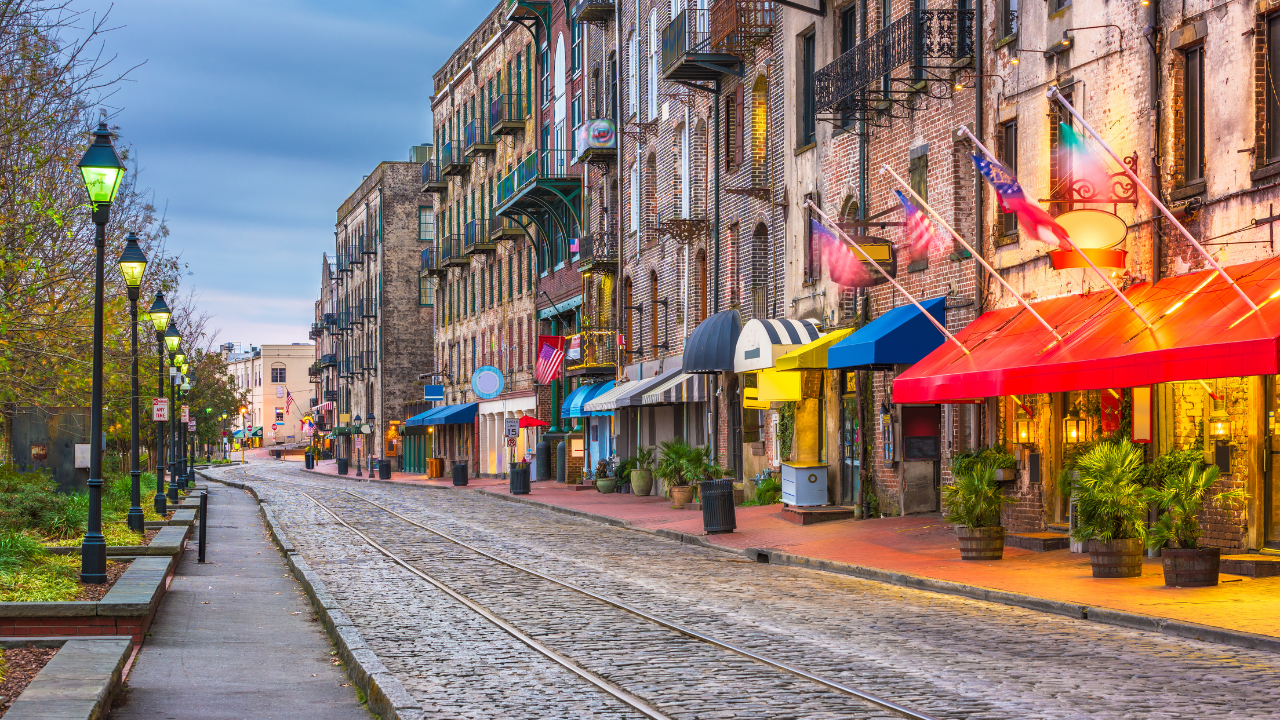 A cobblestone street lined with historic buildings, colorful awnings, and streetlamps. Flags hang from some of the buildings. The sky is overcast.