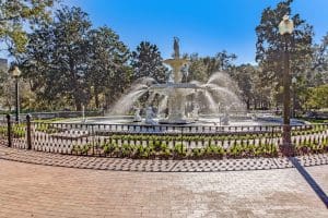 A large, ornate fountain surrounded by a decorative iron fence in a park, with water jets spraying and trees in the background under a clear blue sky.