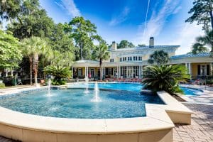 Outdoor pool area with fountains in front of a large house surrounded by trees, under a clear blue sky.