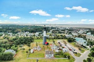 Aerial view of a black-and-white lighthouse with a red roof surrounded by houses, trees, and a parking area under a blue sky.