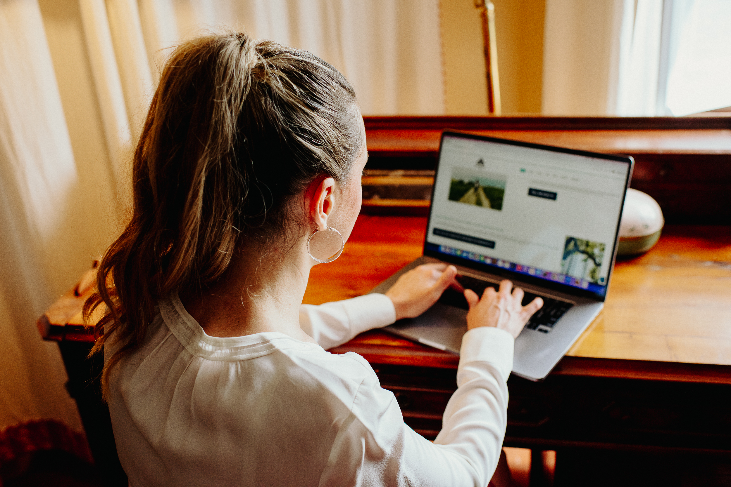 A woman with a ponytail types on a laptop at a wooden desk near a window.