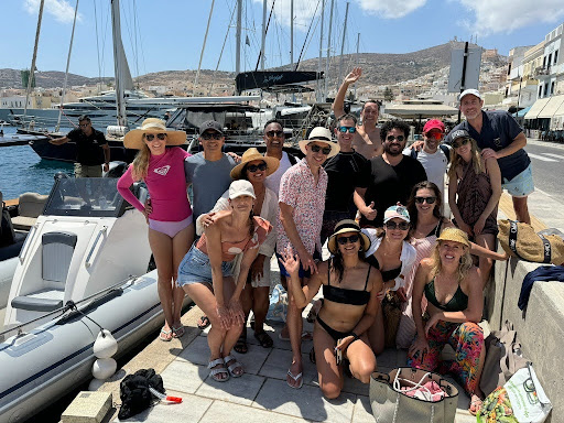A group of people in summer clothing pose and smile by a docked boat in a marina, with sailboats and buildings visible in the background.